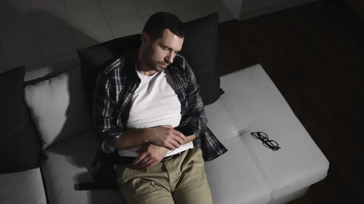 A man wearing a plaid shirt and khaki pants asleep on a gray sofa in a dark room, holding a smartphone with his glasses resting beside him.