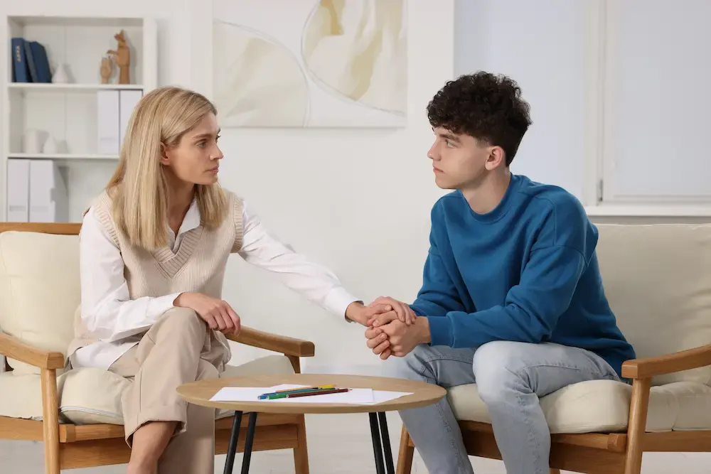 A blonde woman and a young man with curly dark hair sit facing each other in comfortable armchairs in a bright, modern office or counseling setting. The woman reaches across the space between them to gently hold the young man's hands in a supportive gesture. Both have serious expressions and are making direct eye contact. Between them is a small round table with a piece of paper and some colored pencils on it.