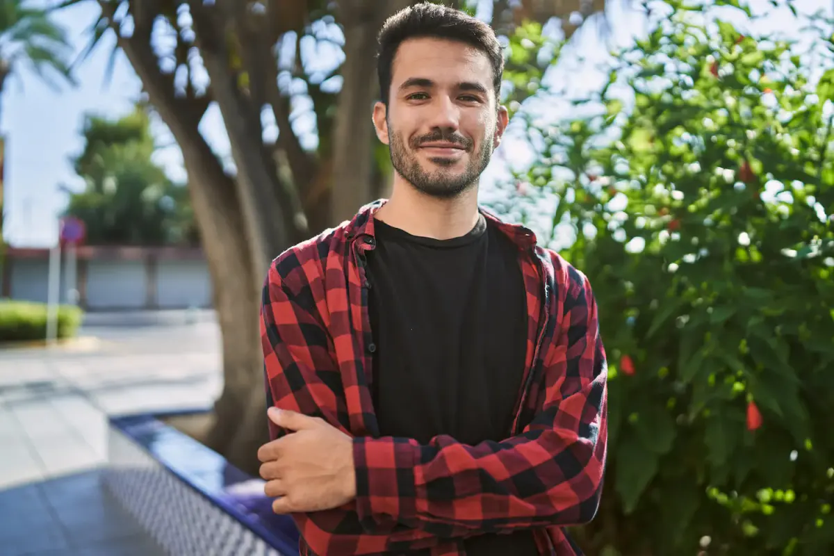 A confident, smiling young Hispanic man standing outdoors, representing strength and successful addiction recovery at More Than Rehab in Texas.