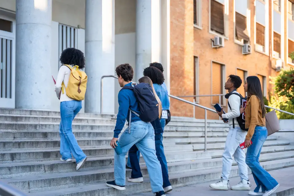 A diverse group of college students with backpacks walking up the steps of a university campus building.