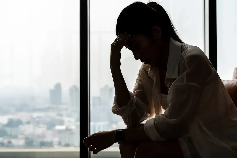 A silhouette of a woman sitting alone, resting her head in her hand in a posture of stress or reflection, positioned against a bright window overlooking a blurred city skyline.
