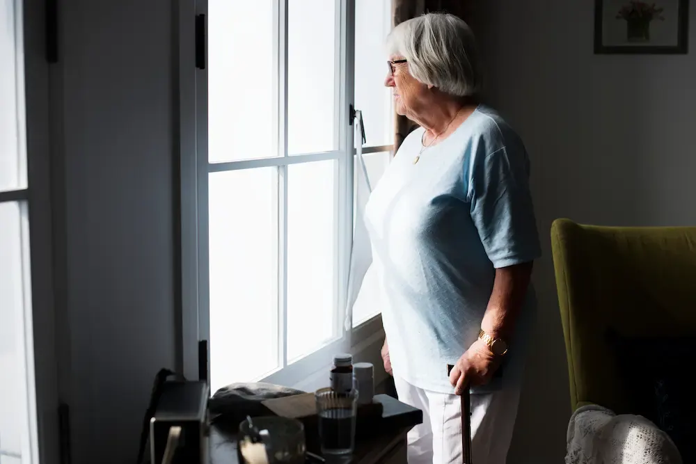 An elderly woman with white hair stands in a dim room, holding a walking cane and gazing out a bright window. On a nearby side table, there is a clear glass of water and several prescription pill bottles, highlighting the "unseen" struggle of medication dependency and isolation among seniors.