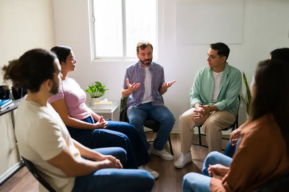 A group of diverse individuals sitting in a circle listening to a speaker during a supportive addiction recovery group therapy session.