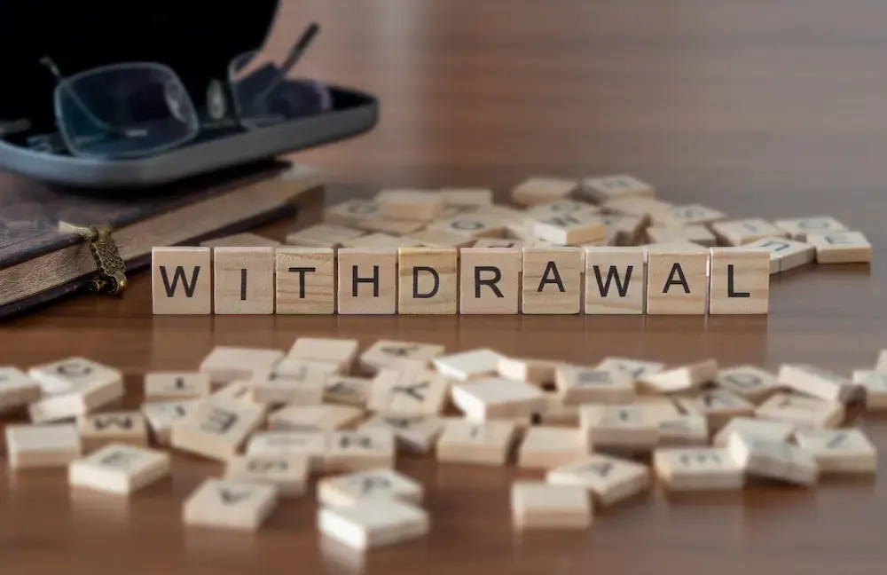 Wooden letter tiles spelling the word 'WITHDRAWAL' lined up in focus on a desk, surrounded by scattered tiles, introducing the concept of drug and alcohol detox.