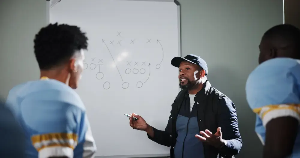 A football coach actively instructing his players in front of a whiteboard with drawn plays.
