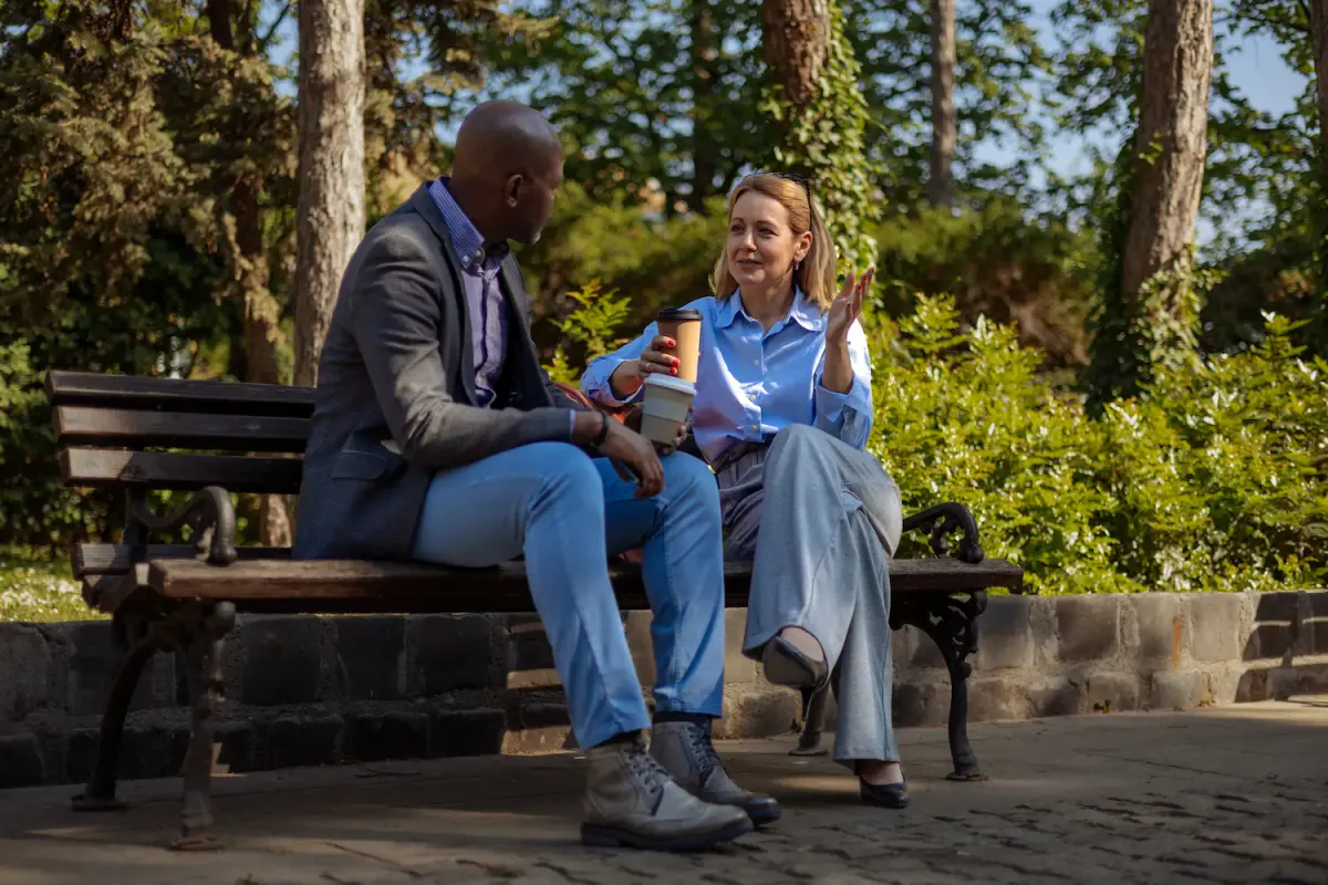 A man and a woman sit together on a sunny park bench, holding coffee cups and engaging in an earnest conversation. The image represents the importance of staying connected to a support network, such as checking in with a sponsor or a sober peer, during the high-risk summer months.